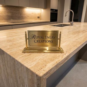 May include: A modern kitchen island with a light brown marble countertop. A brass sign with the text "Royal Inlay Creations" is on the countertop. The kitchen features a stainless steel sink and faucet.
