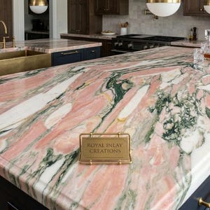 May include: A kitchen island featuring a pink, white, and green marble countertop. The island has dark blue cabinets with gold handles. The countertop has a polished finish and a sign that reads "Royal Inlay Creations".