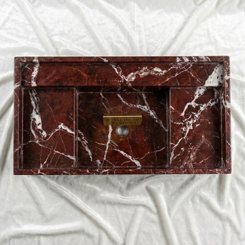 Puede incluir: Un lavabo rectangular de m&aacute;rmol rojo con vetas blancas. El lavabo tiene un orificio para grifo incorporado y varios compartimentos. El texto "ROYAL INLAY CREATIONS" est&aacute; en una placa dorada en la parte delantera del lavabo.