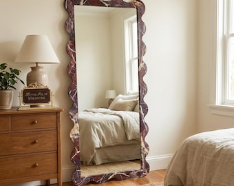 May include: A full-length mirror with a wavy, decorative frame. The frame has a marbled appearance with shades of burgundy, brown, and cream. The mirror reflects a bedroom scene with a bed, dresser, and window. The mirror is a decorative home decor item.