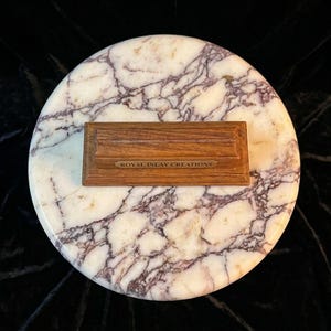 May include: A rectangular wooden display stand with a horizontal groove and a small brass plaque reading ROYAL INLAY CREATIONS, centred on a circular white marble base featuring prominent purple and grey veining throughout the stone surface.