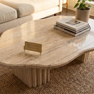 May include: A beige travertine coffee table with an irregular, organic-shaped top and a fluted, cylindrical base. A small gold-toned sign on the surface reads Royal Inlay Creations. The stone features natural veining and a polished finish.