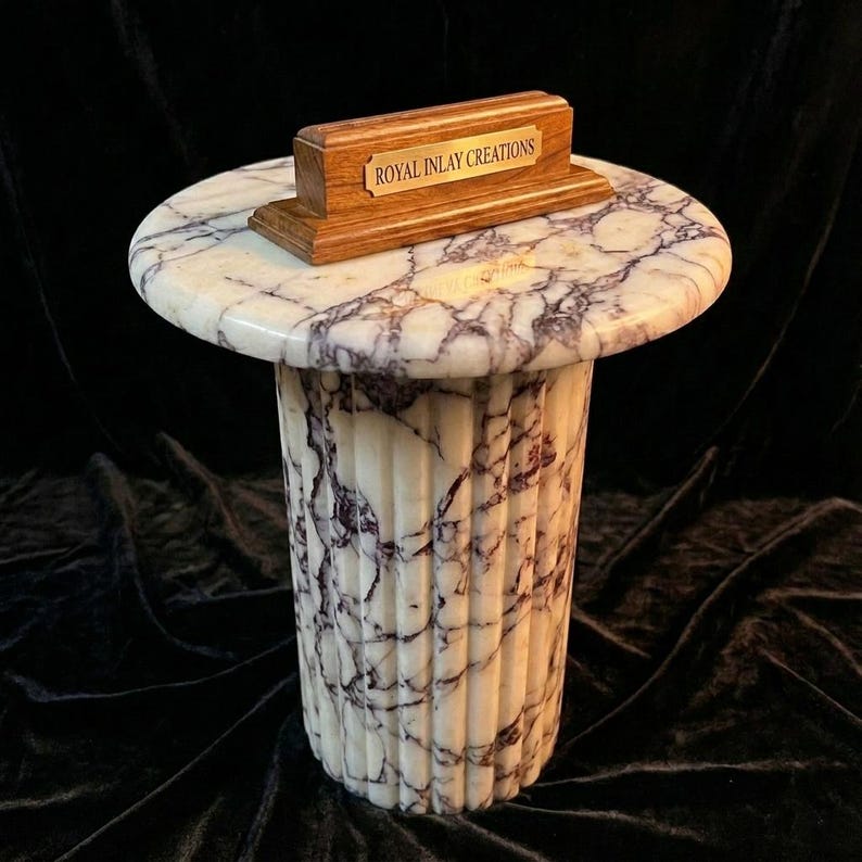 May include: A cylindrical pedestal table featuring a white and purple veined marble top and base with a fluted column design. A rectangular wooden stand sits on the tabletop, displaying a gold-toned metal plaque engraved with ROYAL INLAY CREATIONS.