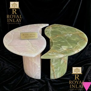 May include: Two onyx side tables, one pink and one green, with a unique, organic shape. Each table has a curved top and a cylindrical base. The tables are from Royal Inlay Creations.