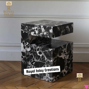 May include: A black and white marble side table with a unique, geometric design. The table features a cube shape with a section removed, creating an open space. The marble has a striking veining pattern. The text "Royal Inlay Creations" is visible.