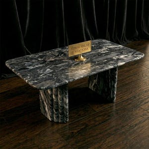 May include: A rectangular marble coffee table with a grey and white pattern. The table has two thick, curved legs and a small gold sign that reads "ROYAL INLAY CREATIONS". The table is on a dark wood floor.