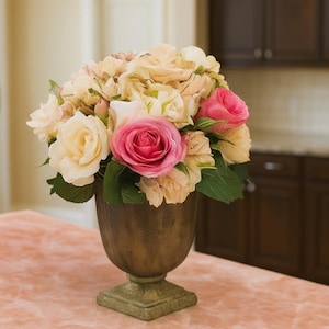 Puede incluir: Un arreglo floral con rosas crema y rosas en una urna de color bronce. La urna tiene una base cuadrada y una superficie texturizada. El arreglo est&aacute; sobre una superficie rosa, con una cocina al fondo.