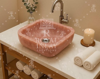 Può includere: Un lavabo quadrato in pietra rosa con un rubinetto in nichel spazzolato. Il lavabo è su un piano di lavoro bianco con una base in legno. Sono presenti anche asciugamani bianchi arrotolati e una candela accesa, creando un'atmosfera da spa.
