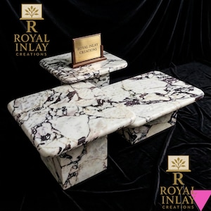 May include: Two marble tables with a white and purple veined pattern. The larger table has a rectangular top and a square base. The smaller table has a square top and a square base. A sign that reads "Royal Inlay Creations" is on the smaller table.