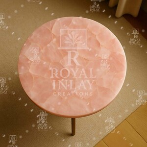May include: A round pink table with a natural stone top. The table is made of rose quartz and has a unique, textured surface. The table is supported by a single, gold-colored leg.