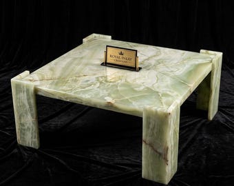 Puede incluir: Una mesa de centro cuadrada de ónix verde con una superficie pulida y cuatro patas robustas. La mesa presenta un patrón de vetas natural y un pequeño letrero dorado que dice "ROYAL INLAY CREATIONS". La mesa está sobre un fondo negro.