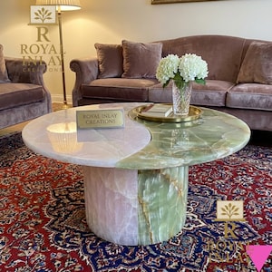 May include: A two-tone coffee table with a pink and green marble top. The table has a round shape with a yin-yang design. The base is cylindrical and made of matching marble. The table is in a living room setting with a sofa and rug.