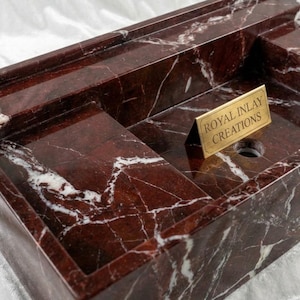Puede incluir: Un lavabo rectangular de m&aacute;rmol rojo con vetas blancas. El lavabo tiene un estante incorporado y un orificio de drenaje. Un peque&ntilde;o letrero dorado que dice "ROYAL INLAY CREATIONS" est&aacute; colocado en el lavabo.