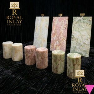May include: A display of stone pedestals and panels in various colors. The cylindrical pedestals are beige, pink, and green. The rectangular panels are white, pink, and green with veining. The Royal Inlay Creations logo is visible.