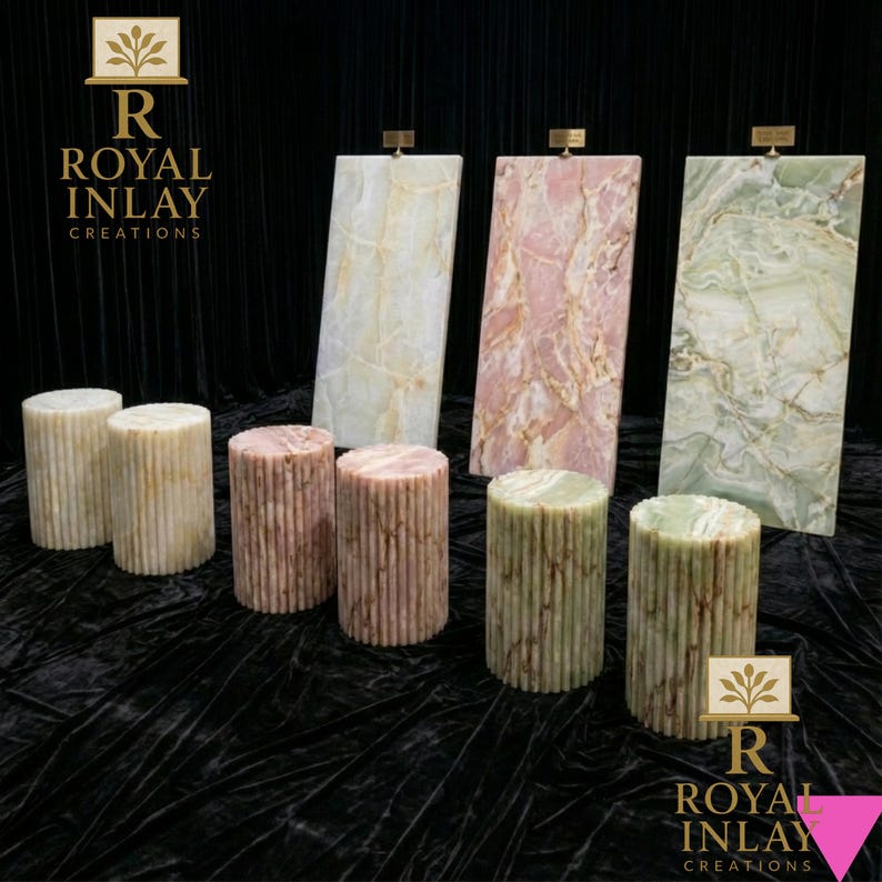 May include: A display of stone pedestals and panels in various colors. The cylindrical pedestals are beige, pink, and green. The rectangular panels are white, pink, and green with veining. The Royal Inlay Creations logo is visible.