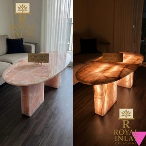 Op de afbeelding: Roze onyx ovale salontafel met een gepolijst oppervlak en rechthoekige voet. De tafel is verlicht in de tweede afbeelding, wat de doorschijnende kwaliteiten laat zien. De tafel heeft de tekst "ROYAL INLAY CREATIONS".