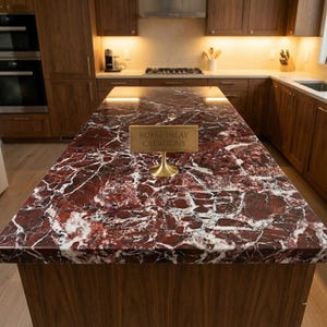 May include: A kitchen island with a red and white marble countertop. The countertop has a unique veining pattern. The island is made of dark wood. A gold sign that reads "Royal Inlay Creations" is on the countertop.