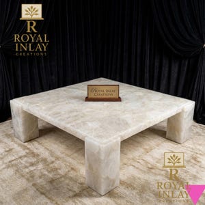 May include: A large, square coffee table made of a light-coloured, veined stone. The table has a solid, blocky base and a smooth, polished surface. The table is labelled with the text "Royal Inlay Creations".