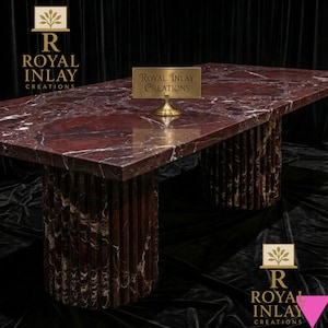 Puede incluir: Mesa rectangular de mármol rojo con vetas blancas. Tiene dos patas cilíndricas estriadas. Un pequeño letrero dorado con el texto "Royal Inlay Creations" está sobre la mesa. Una pieza destacada para cualquier comedor o sala de estar. Mide unos 200 cm de largo.