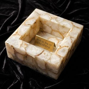 May include: A square bowl constructed from translucent white and beige agate stone slices. The interior features a small, rectangular gold-toned metal plaque with the text ROYAL INLAY CREATIONS engraved in a dark font on its surface.