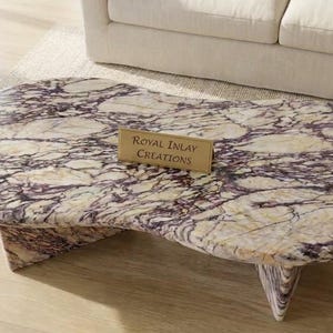 Puede incluir: Una mesa de centro con un tablero de forma orgánica irregular hecho de piedra pulida con un patrón veteado en color crema y morado oscuro. La mesa se apoya en una base de piedra a juego y tiene una pequeña placa dorada que dice Royal Inlay Creations.