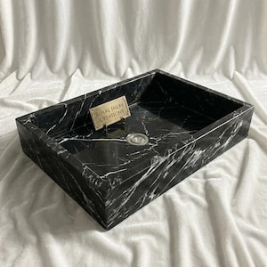Könnte beinhalten: Ein rechteckiges Waschbecken aus schwarzem Marmor mit weißen Adern. Das Waschbecken hat eine polierte Oberfläche und einen Edelstahl-Abfluss. Ein kleines goldfarbenes Schild mit dem Text "ROYAL INLAY CREATIONS" ist angebracht.