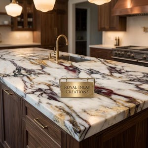 May include: A large kitchen island featuring a white marble countertop with prominent black and deep red veining. A gold-toned metal sign with the text Royal Inlay Creations sits on the surface. The island includes a gold faucet and sink.
