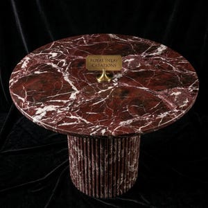 Puede incluir: Una mesa redonda de m&aacute;rmol con una tapa veteada en rojo oscuro y blanco. La base es cil&iacute;ndrica, hecha de piezas verticales de m&aacute;rmol rojo y blanco. Un peque&ntilde;o letrero de lat&oacute;n dice "ROYAL INLAY CREATIONS".