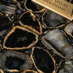 May include: Close-up of a decorative surface featuring black and gray agate stone slices with gold-colored edging. The stone slices are irregularly shaped and create a mosaic-like pattern. A gold-colored sign with the text "INLAY CREATIONS" is in the upper right corner.