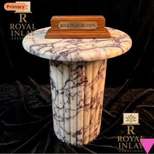 May include: A round side table featuring a white marble top with purple veining and a matching fluted cylindrical base. A rectangular wooden stand with a brass plaque reading ROYAL INLAY CREATIONS sits on the table surface.