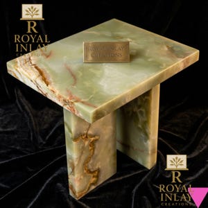 Puede incluir: Una mesa auxiliar cuadrada de ónix verde con una placa rectangular de latón que dice "Royal Inlay Creations". La mesa tiene una base de dos patas y una superficie pulida con vetas marrones y rojas. La mesa es una pieza de mobiliario decorativa.