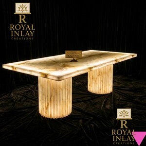 May include: A rectangular, illuminated onyx table with two cylindrical, fluted legs. The table's surface has a marble-like pattern. The table is branded with the text "Royal Inlay Creations".
