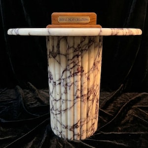May include: A white marble pedestal table with dark purple veining and a fluted cylindrical base. A rectangular wooden block sits on the centre of the tabletop, featuring a brass-coloured metal plaque with the text ROYAL INLAY CREATIONS.