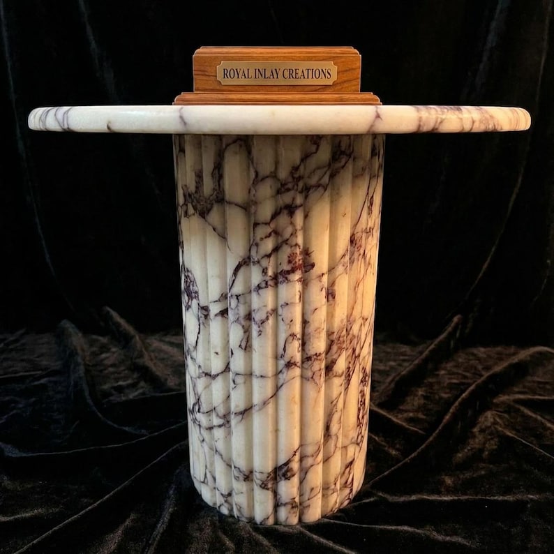 May include: A white marble pedestal table with dark purple veining and a fluted cylindrical base. A rectangular wooden block sits on the centre of the tabletop, featuring a brass-coloured metal plaque with the text ROYAL INLAY CREATIONS.