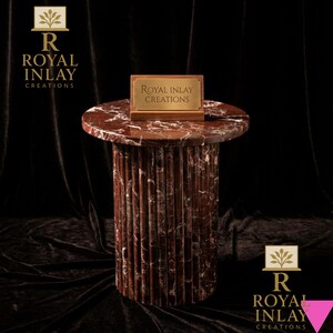 May include: A red marble side table with a round top and a fluted column base. A small rectangular plaque with the text "Royal Inlay Creations" sits on top. The table is set against a dark background.