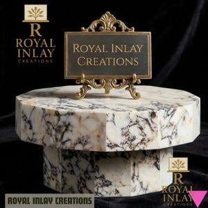 Può includere: Un espositore in marmo dal design sfaccettato. L'espositore presenta un cartello rettangolare con la scritta "ROYAL INLAY CREATIONS" in lettere dorate, sostenuto da un cavalletto dorato ornato. L'espositore è bianco con venature marroni e grigie.