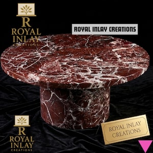 Puede incluir: Una mesa redonda de mármol rojo con vetas blancas. Tiene una base cilíndrica y una superficie pulida. El texto "ROYAL INLAY CREATIONS" se muestra en la mesa y en una placa dorada. El fondo es negro.
