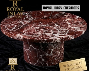 May include: A round, red marble table with white veining. The table has a cylindrical base and a polished surface. The text "ROYAL INLAY CREATIONS" is displayed on the table and on a gold plaque. The table is set against a black background.