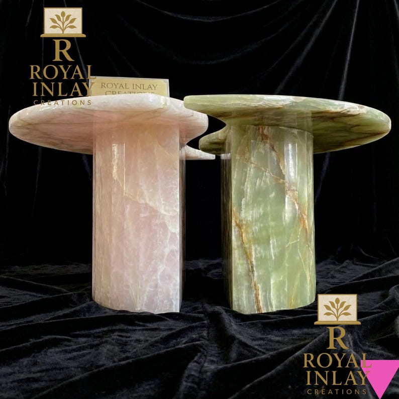 May include: Two stone side tables. One is a light pink color, and the other is green. Both tables have a round top with a unique shape and a cylindrical base. The tables are made of a polished stone material. The text "ROYAL INLAY CREATIONS" is visible.