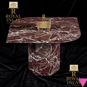 May include: A red marble table with white veining, featuring a unique, curved tabletop. The table has a geometric base and a small gold sign that reads "Royal Inlay Creations". The table is set against a dark background.