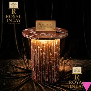 May include: A round, marble-topped side table with a cylindrical base. The table has a reddish-brown color with white veining. The base is illuminated from within, creating a warm glow. The table features a small sign that reads "Royal Inlay Creations".