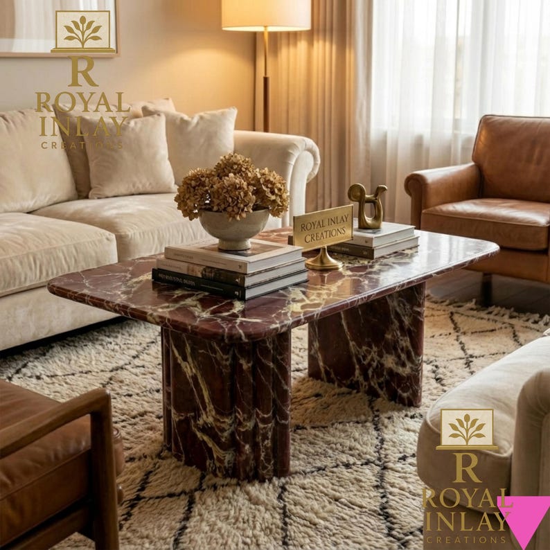 May include: A rectangular coffee table with a red marble top and matching base. The table is in a living room setting with a beige sofa, leather chairs, and a patterned rug. A vase of dried flowers and books are on the table.