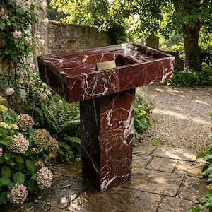 Puede incluir: Un lavabo exterior de m&aacute;rmol rojo con vetas blancas, colocado sobre un pedestal a juego. El lavabo tiene una cubeta rectangular con una peque&ntilde;a abertura rectangular. El lavabo est&aacute; rodeado de exuberante vegetaci&oacute;n y un camino de piedra. Una peque&ntilde;a placa dorada dice "Royal Inlay Creations."