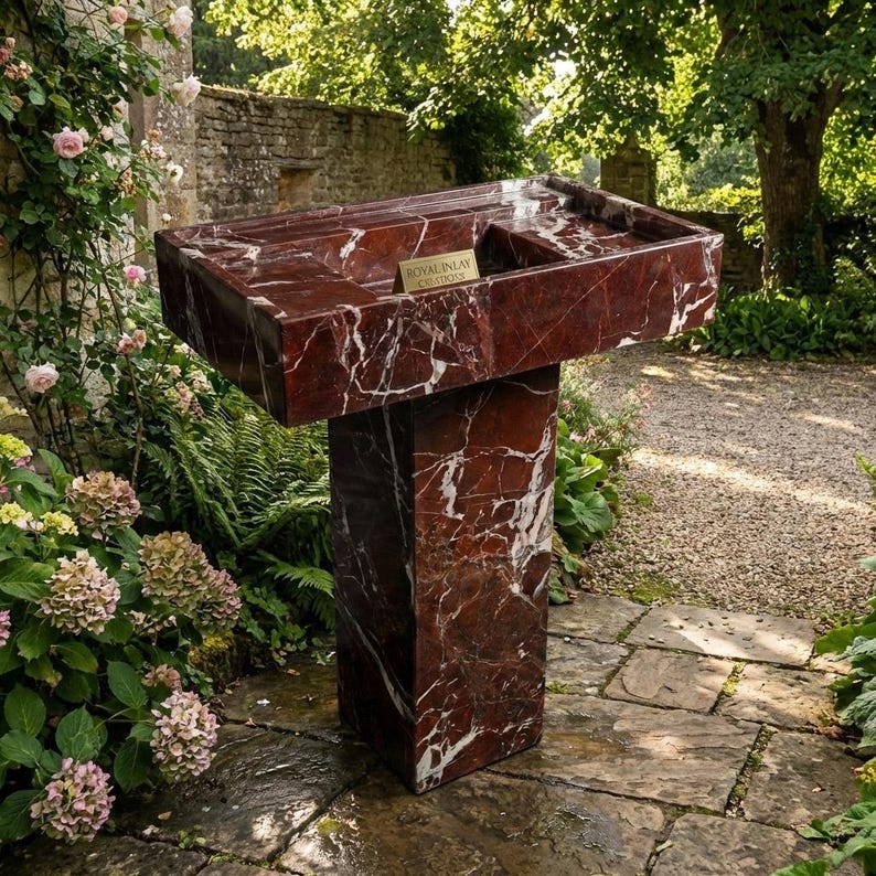 Puede incluir: Un lavabo exterior de m&aacute;rmol rojo con vetas blancas, colocado sobre un pedestal a juego. El lavabo tiene una cubeta rectangular con una peque&ntilde;a abertura rectangular. El lavabo est&aacute; rodeado de exuberante vegetaci&oacute;n y un camino de piedra. Una peque&ntilde;a placa dorada dice "Royal Inlay Creations."