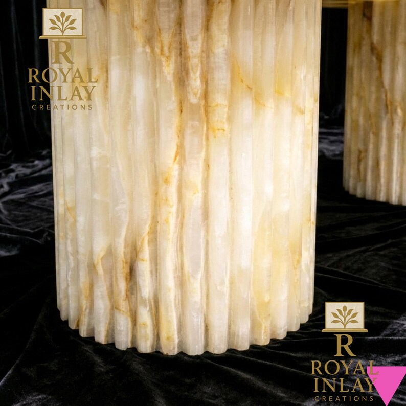 May include: Close-up of a cylindrical, fluted table base made of translucent, cream-colored onyx with golden veining. The base has a scalloped edge. The logo "ROYAL INLAY CREATIONS" is visible.