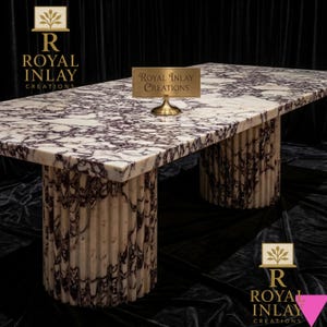 Puede incluir: Una mesa rectangular de mármol con una parte superior con estampado blanco y morado. La mesa tiene dos bases cilíndricas con detalles de nervaduras verticales. Un pequeño letrero dorado con el texto "Royal Inlay Creations" está sobre la mesa. La mesa está frente a un fondo negro.