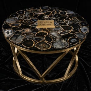 May include: Round coffee table with a black and gray agate stone top and a gold metal base. The tabletop is made of polished agate stones with gold-colored edges. The table has a geometric base and the text "ROYAL INLAY CREATIONS".