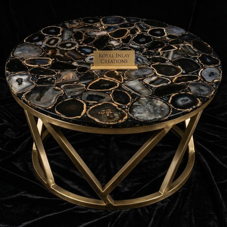 May include: Round coffee table with a black and gray agate stone top and a gold metal base. The tabletop is made of polished agate stones with gold-colored edges. The table has a geometric base and the text "ROYAL INLAY CREATIONS".