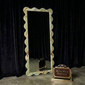 May include: A tall, rectangular mirror with a wavy, light green and brown onyx frame. The mirror is standing upright against a black curtain backdrop. A small, decorative sign with the text "Royal Inlay Creations" is in front of the mirror.