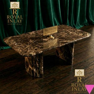 Puede incluir: Una mesa de centro rectangular de mármol con vetas marrones y beige. La mesa tiene una superficie pulida y dos patas gruesas y texturizadas. Un pequeño letrero dorado dice "Royal Inlay Creations". El logotipo "Royal Inlay Creations" también es visible.